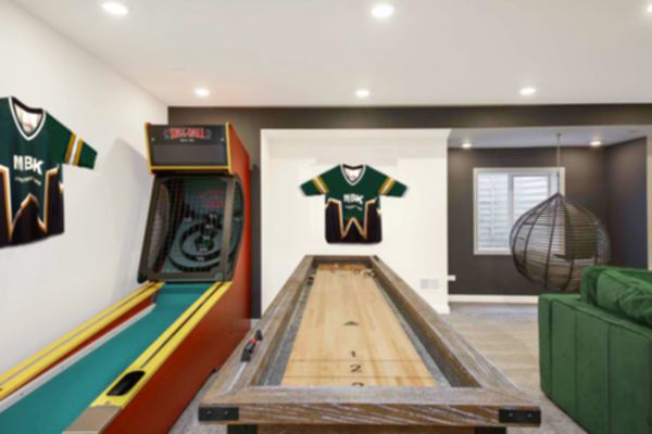 An image of a basement remodel with a skee ball game and a curling game, there are 2 "MBK Constructors Hockey Jersey's hanging on the wall, it has bright recessed lighting