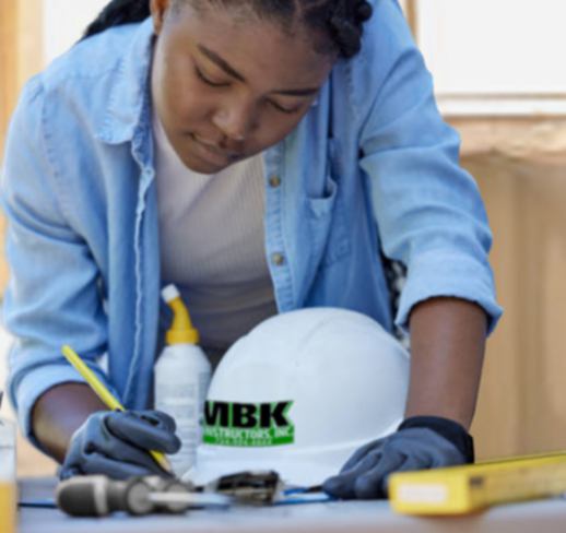 An image of a Black Female Carpenter at work. She is leaning over a table checking off items on a clipboard, she is wearing a blue button down work shirt with a white t-shirt, she has a focused and satisfied look on her face.